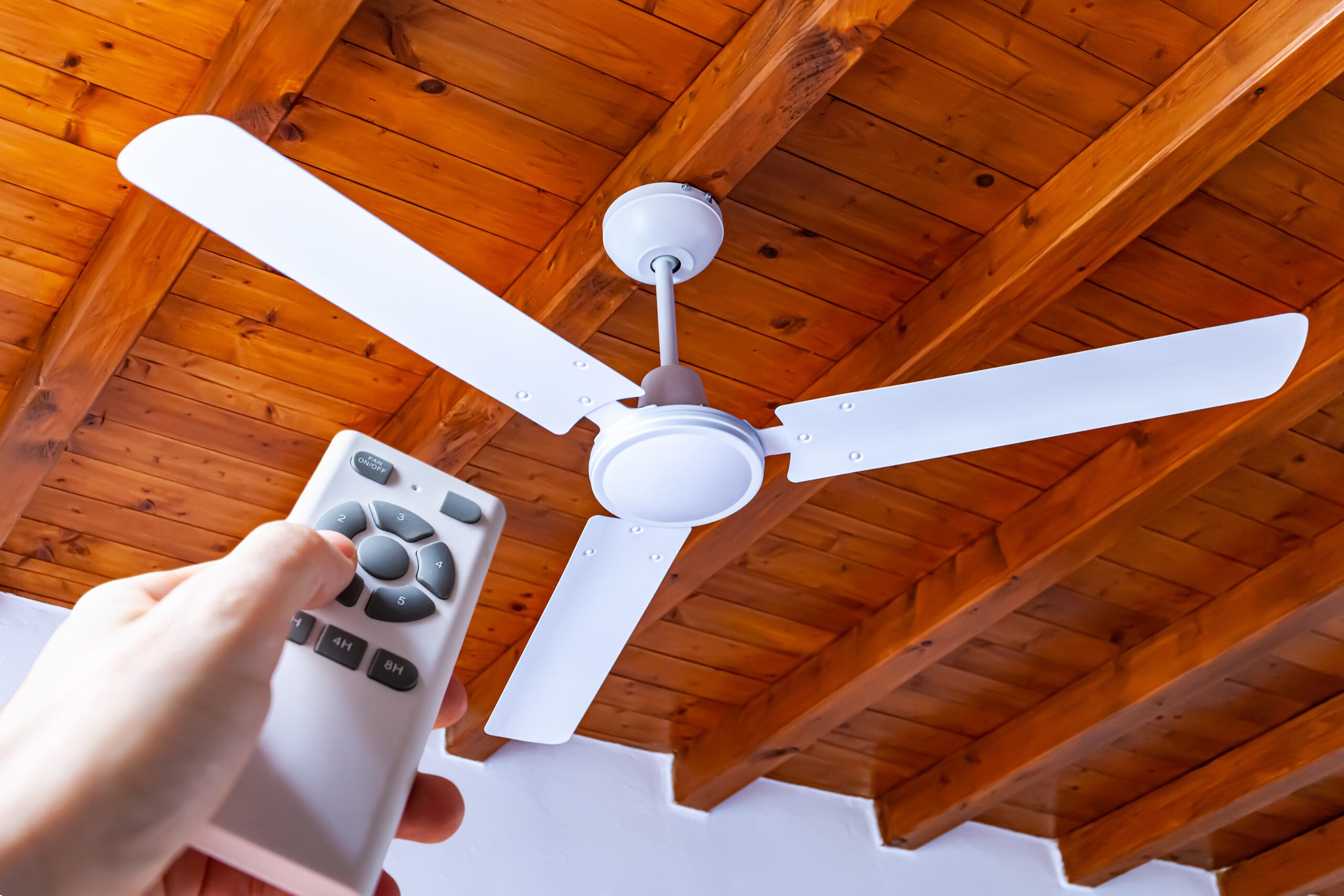 A man uses a remote control to turn on a white ceiling fan mounted in a house with wooden ceilings.