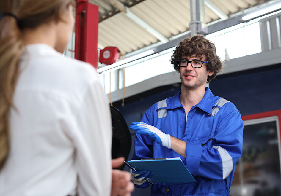 Man technician car mechanical in uniform showing car maintenance service report on clipboard at repair garage station. Auto mechanic give customer discussion on her vehicle repairs problems condition